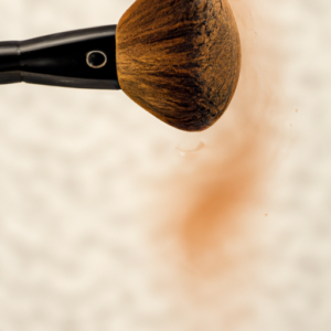 A closeup of a makeup brush making a circular motion on a face with a neutral-colored background.