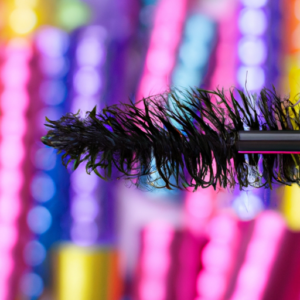 A close up of a mascara brush against a background of colorful and vibrant eyelashes.