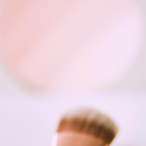 Soft pink and white gradient background, with a blurred reflection of a makeup brush in the foreground.